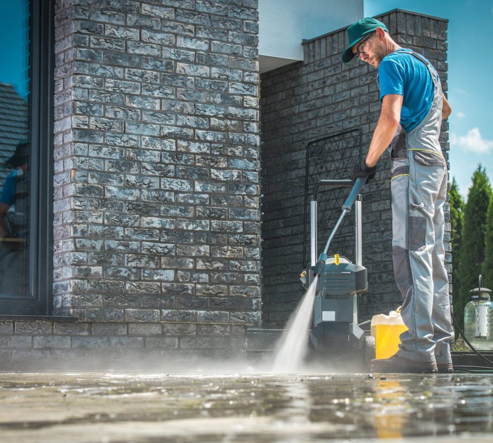 Pressure Washer Cleaning Pressure Washer Cleaning in Front of the House. Caucasian Men in His 30s Washing Concrete Bricks Driveway in Sunny Summer Day. Cleaning Around the House Concept.